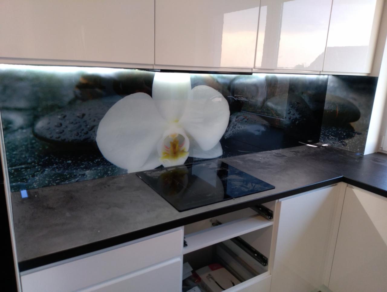 Szklany panel kuchenny z motywem białej orchidei na tle kamieni, podświetlony listwą LED, zamontowany nad płytą indukcyjną w nowoczesnej kuchni z białymi szafkami.