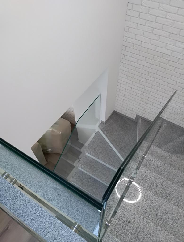 Schody wykończone szarym kamieniem z balustradą ze szkła hartowanego, widok z góry, ściana z białej cegły w tle.