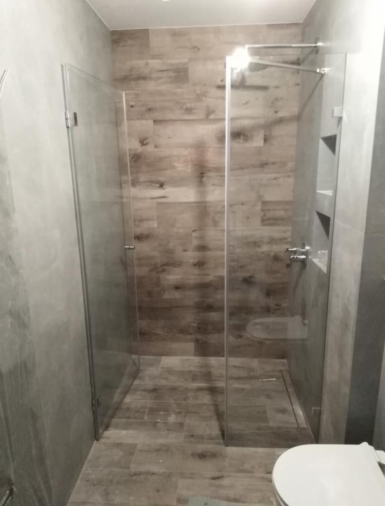 Kabina prysznicowa ze szklanymi drzwiami, wykończona płytkami imitującymi drewno, z widocznymi półkami w ścianie.