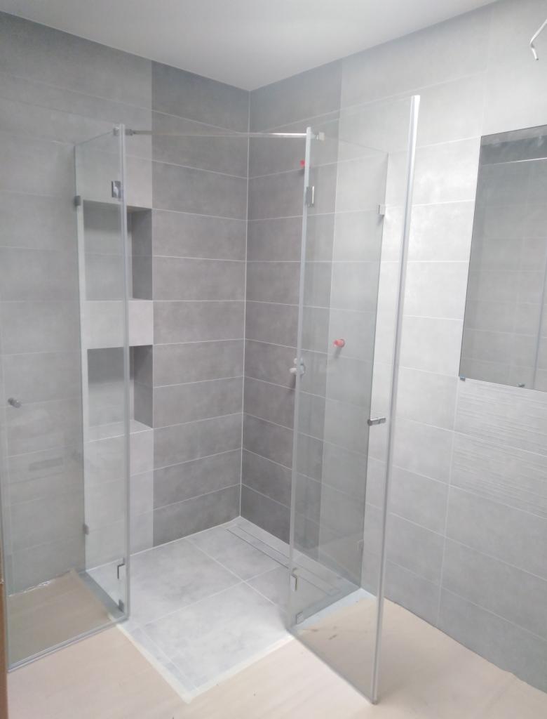 Szklana kabina prysznicowa z dwoma ściankami i drzwiami, zamontowana w łazience wyłożonej szarymi płytkami, widoczne listwy odpływowe w podłodze.