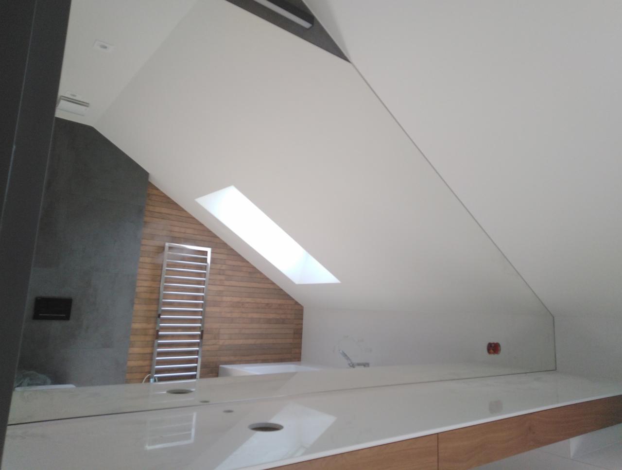 Nowoczesna łazienka na poddaszu z oknem dachowym, widoczna umywalka z blatem z jasnego marmuru i drewniane szafki pod spodem, grzejnik drabinkowy na ścianie z drewnianą okładziną. Nietypowy układ...