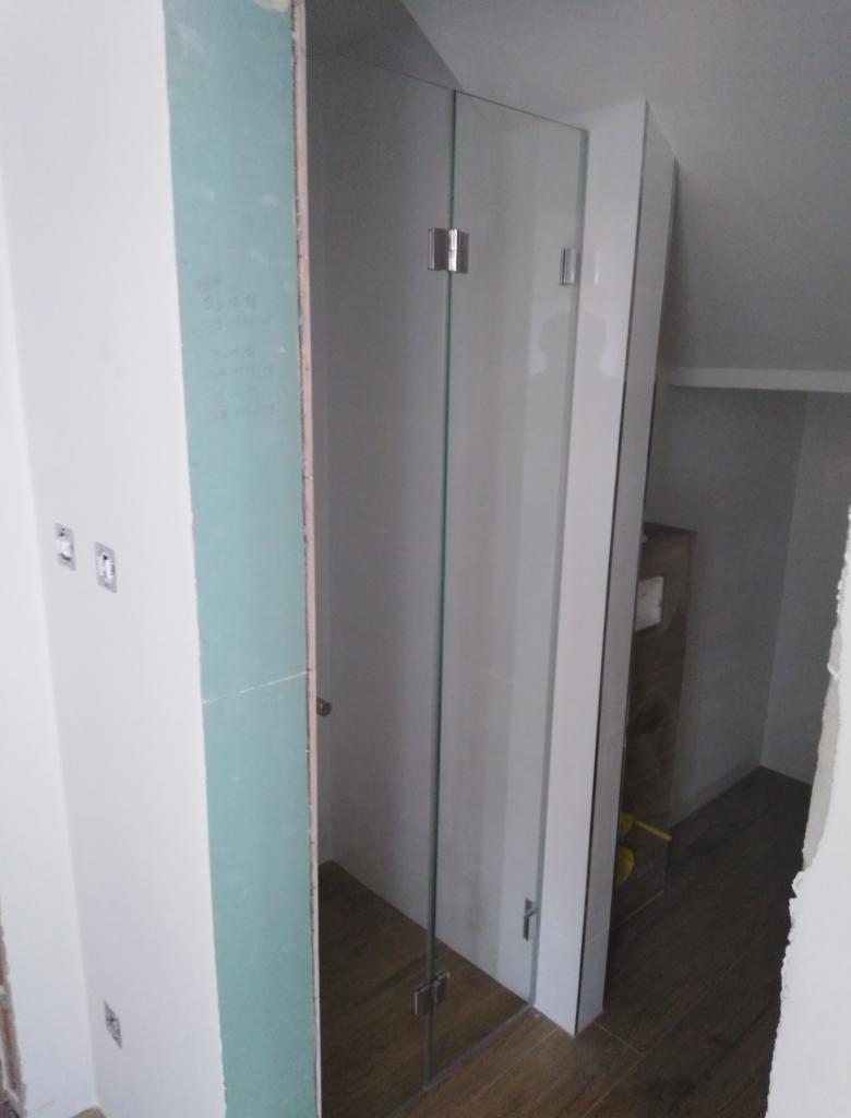 Montaż kabiny prysznicowej ze szklanymi drzwiami, widoczne zawiasy i fragment ściany z płyt gipsowo-kartonowych przed wykończeniem w łazience na poddaszu.