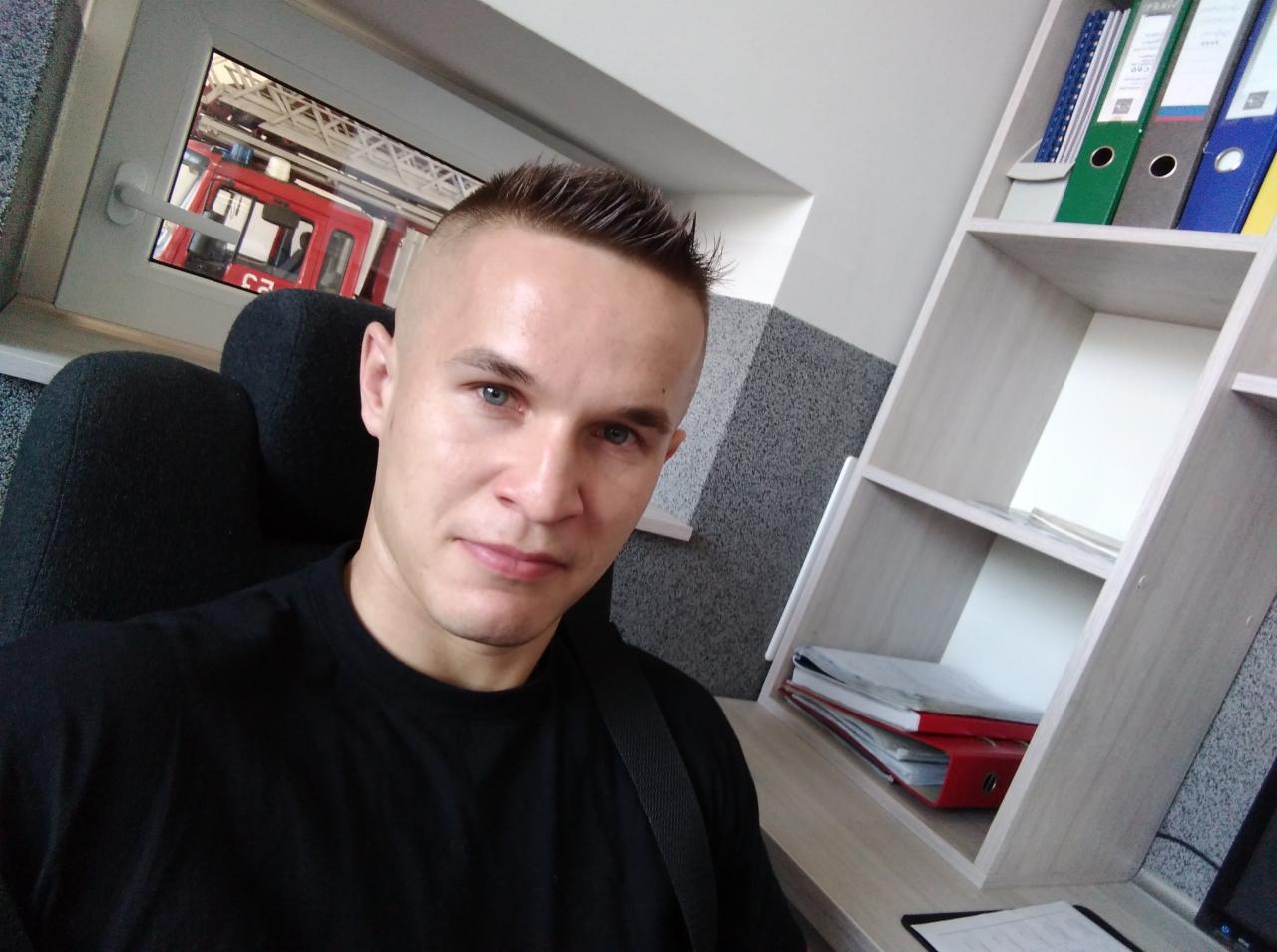 Mężczyzna w czarnej koszulce siedzi na krześle; w tle widać wóz strażacki za oknem i regał z segregatorami.