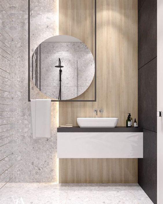 Nowoczesna łazienka z umywalką nablatową na szafce wiszącej, okrągłym lustrem w czarnej ramie odbijającym prysznic, wykończona płytkami imitującymi kamień i drewno.