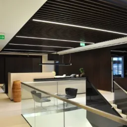 Nowoczesne biuro z drewnianym sufitem rastrowym i liniowym oświetleniem LED, widok z antresoli na recepcję z przeszkloną balustradą i schodami.