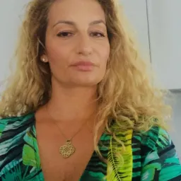 Portret kobiety z blond lokami, w zielonej sukience z motywem liści, złoty naszyjnik z ozdobnym wisiorkiem. Widoczne białe szafki w tle.