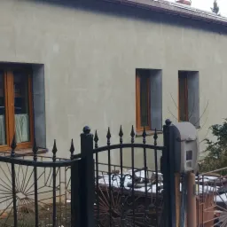 Elewacja domu jednorodzinnego po nałożeniu szarej, surowej gładzi, widoczna zza ozdobnego metalowego ogrodzenia w zimowej scenerii.