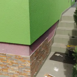 Fragment elewacji budynku z zieloną i różową warstwą oraz dekoracyjną okładziną z płytek imitujących kamień, widoczne krzyżyki dystansowe, obok schody wejściowe i tuja.