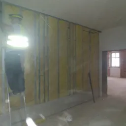 Prace związane z izolacją ścian wełną mineralną w Zawierciu, widoczny pracownik na drabinie w kamizelce odblaskowej i metalowy stelaż pod płyty gipsowo-kartonowe.