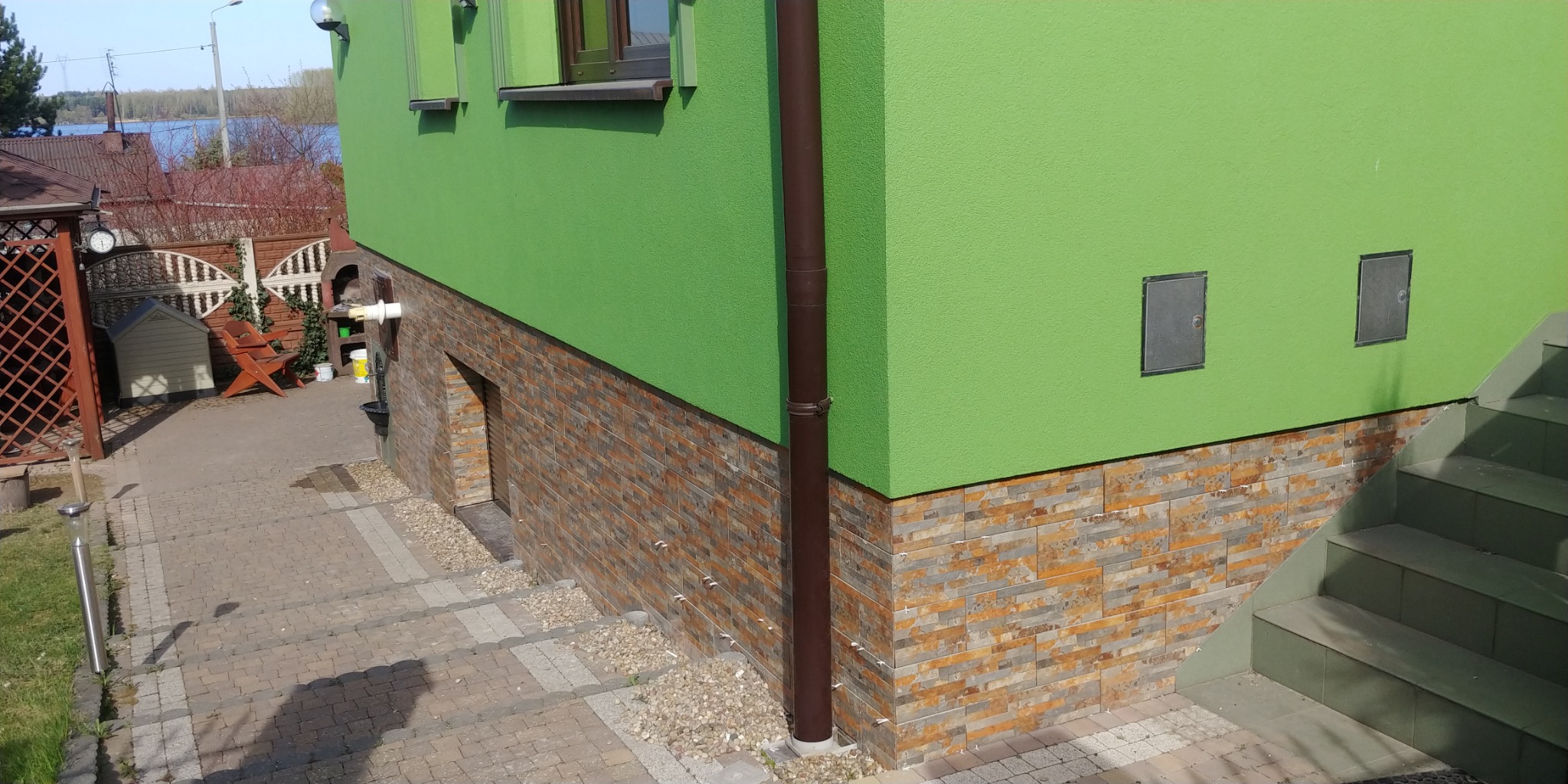 Jasnozielony budynek z elewacją wykończoną płytkami imitującymi kamień, brązowa rynna, małe drzwi wejściowe do piwnicy i schody prowadzące do góry.