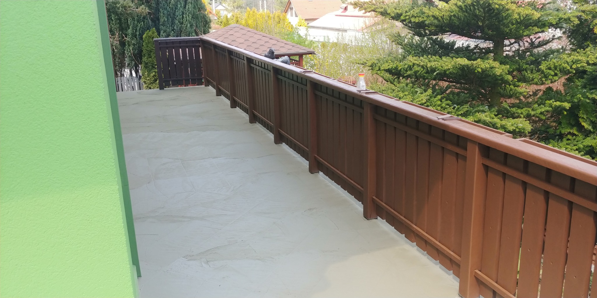 Balkon po nałożeniu szarej wylewki samopoziomującej, z brązową balustradą i widokiem na zieleń w tle. Na balustradzie siedzą dwa gołębie.