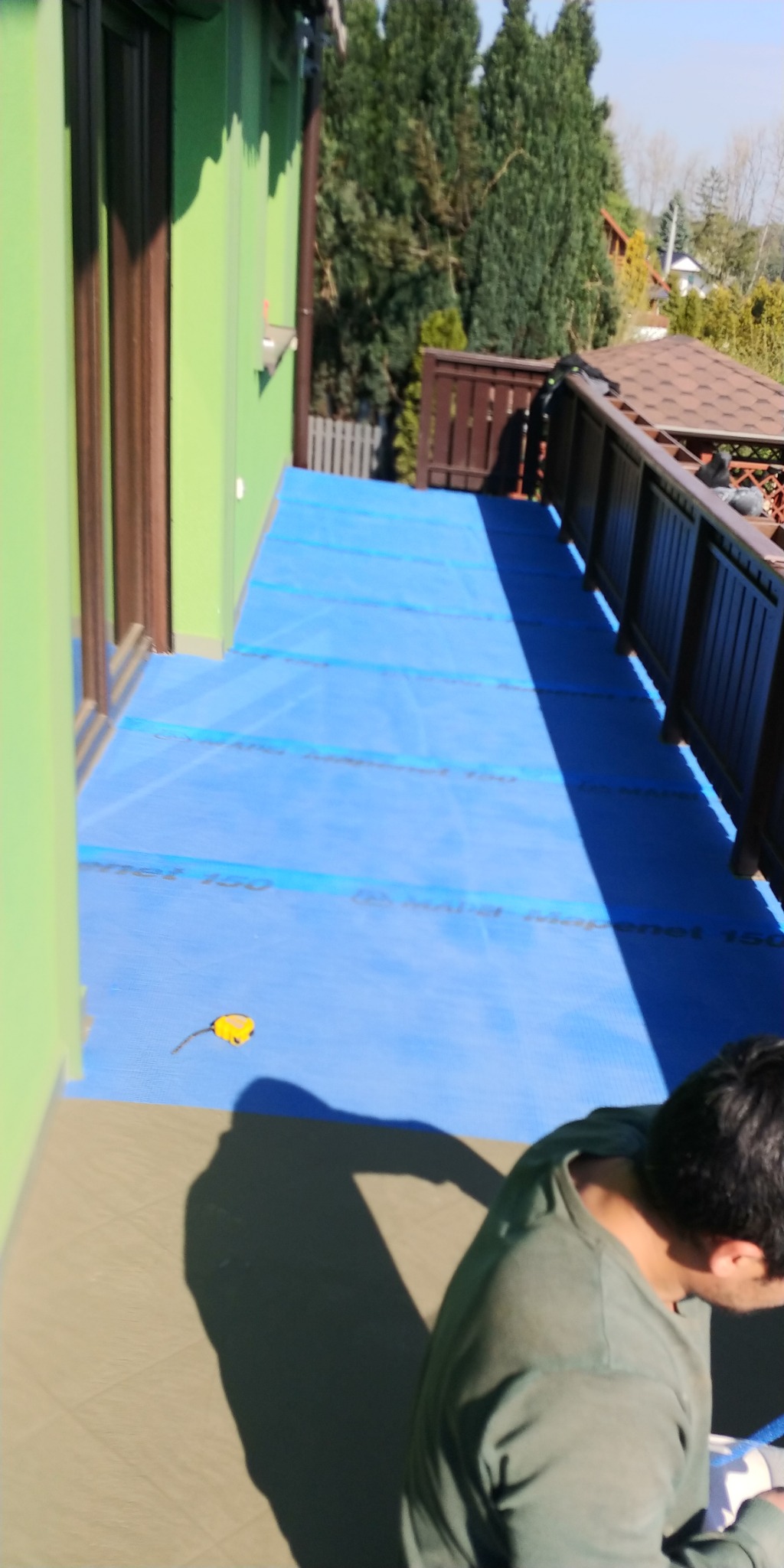 Balkon zabezpieczony niebieską membraną hydroizolacyjną Mapenet 150 przed dalszymi pracami, widoczny zwinięty metr i fragment sylwetki pracownika.