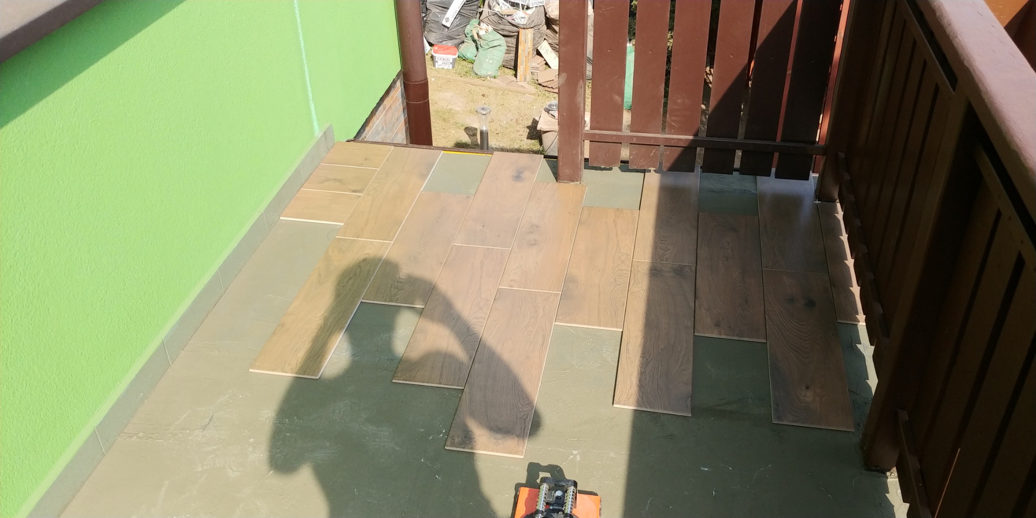 Układanie drewnopodobnych paneli na balkonie, widoczny cień osoby wykonującej prace i narzędzie do docinania paneli.