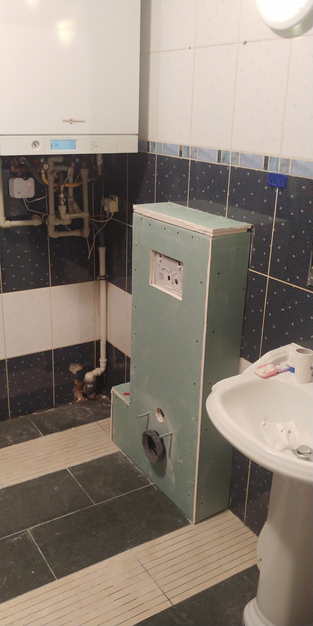 Stelaż podtynkowy obudowany płytami gipsowo-kartonowymi w trakcie montażu w łazience, widoczny otwór na rurę odpływową, grzejnik gazowy na ścianie, umywalka na postumencie.