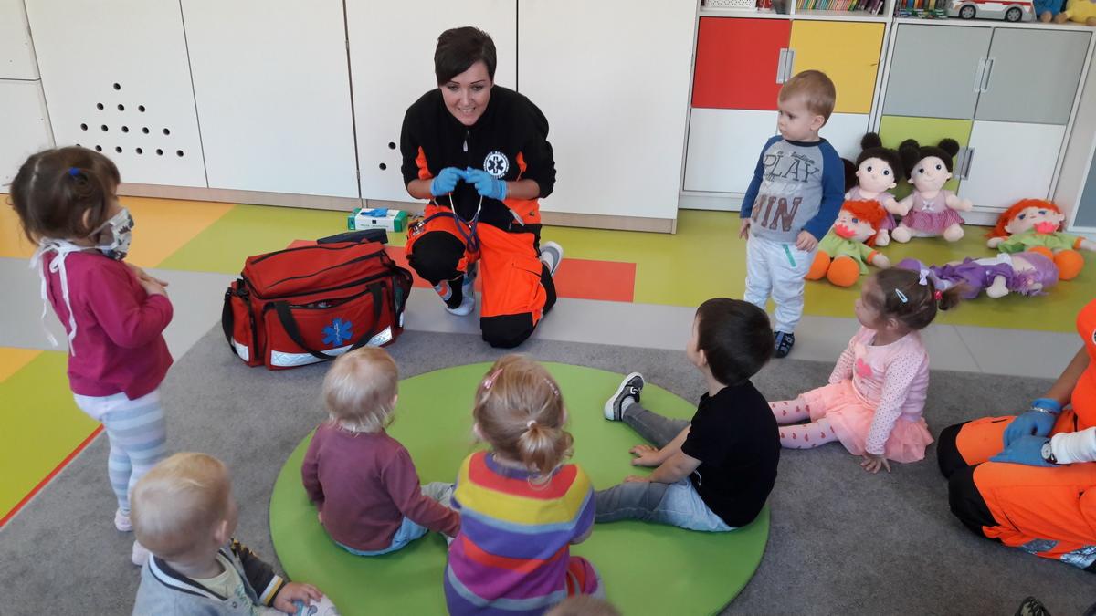 Ratownik medyczny w pomarańczowym uniformie demonstruje dzieciom w przedszkolu użycie stetoskopu, obok czerwona torba medyczna z symbolem gwiazdy życia.