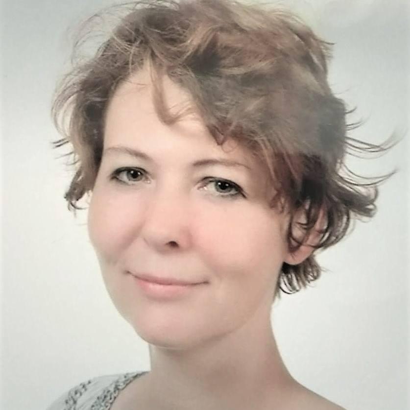 Portret kobiety o krótkich, brązowych włosach, z delikatnym uśmiechem na jasnym tle. Widoczne subtelne zmarszczki mimiczne wokół oczu.