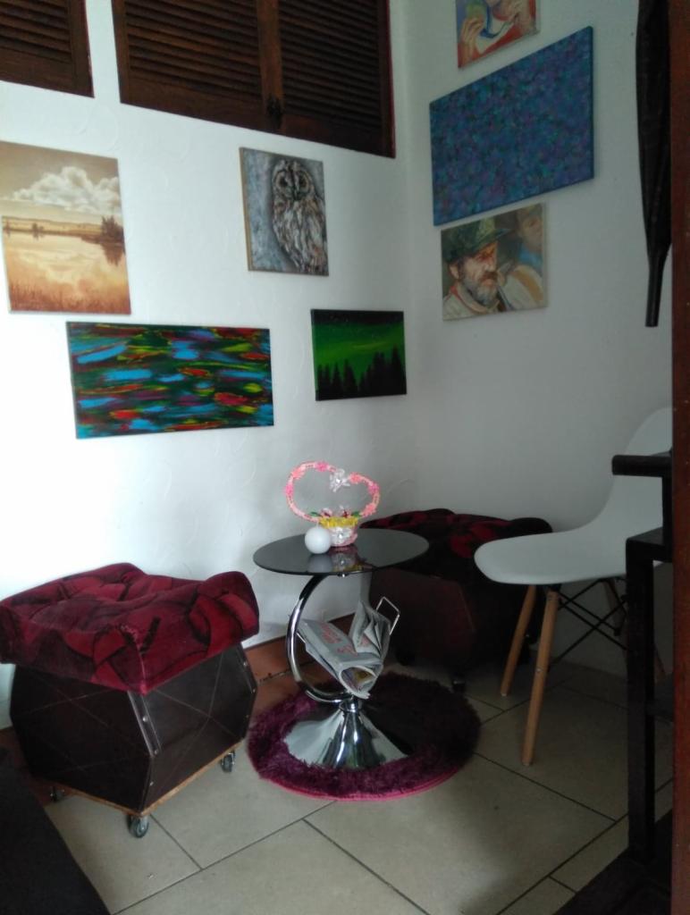 Ściana z różnorodnymi obrazami, od pejzaży po portrety i abstrakcje, uzupełniona stolikiem z dekoracją w kształcie serca i gazetnikiem, obok pufa i krzesło.