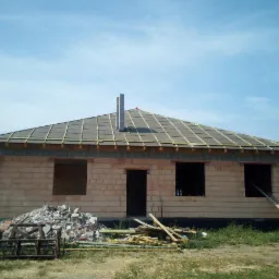 Surowy stan budynku jednorodzinnego z częściowo położonym dachem z dachówki ceramicznej, widoczne kominy i konstrukcja więźby, na pierwszym planie gruz i materiały budowlane.