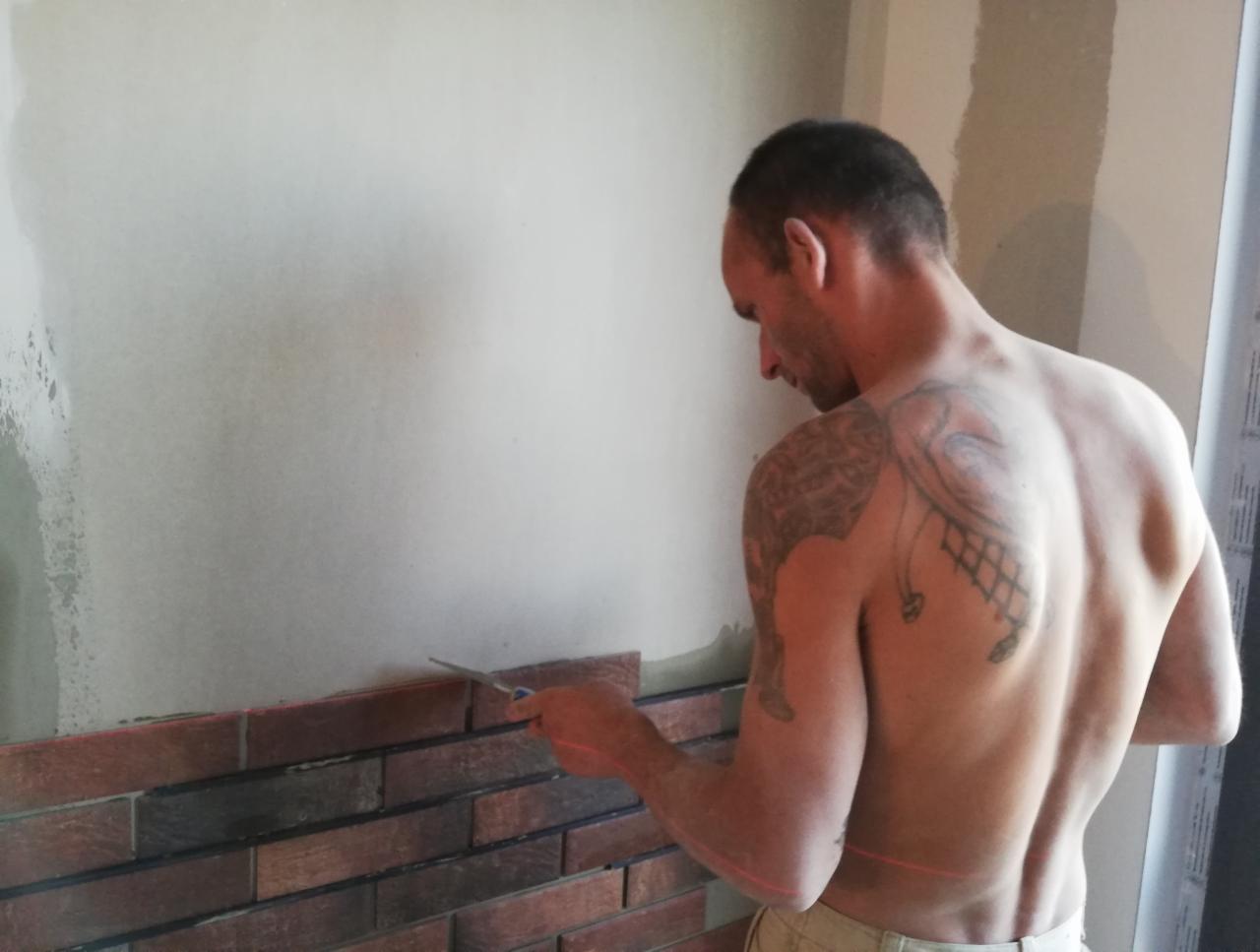 Mężczyzna z tatuażem układa cegiełki na ścianie, używając poziomicy laserowej. Widoczny fragment ściany z surowym wykończeniem i ułożone cegiełki w odcieniach brązu.