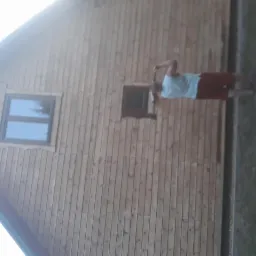 Malowanie drewnianej elewacji domu, widoczna osoba z wałkiem malarskim przy oknie, świeżo pomalowane deski.