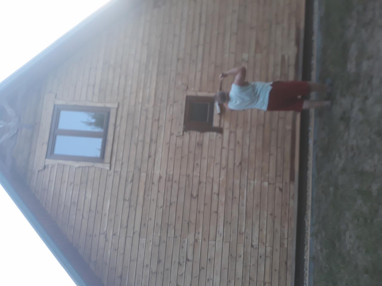 Malowanie drewnianej elewacji domu, widoczna osoba z wałkiem malarskim przy oknie, świeżo pomalowane deski.