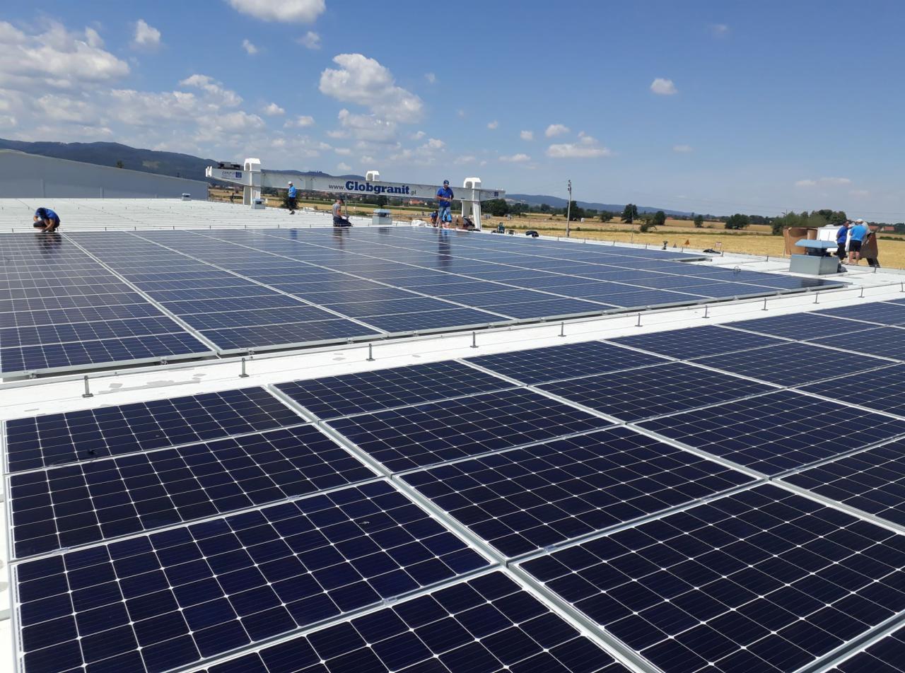 Instalacja paneli słonecznych na dachu przemysłowym z widocznym logo firmy na dachu budynku w tle, błękitne niebo.