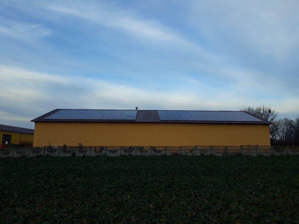 Żółty budynek gospodarczy z panelami słonecznymi na dachu, widoczne na tle pochmurnego nieba i za niskim betonowym ogrodzeniem.