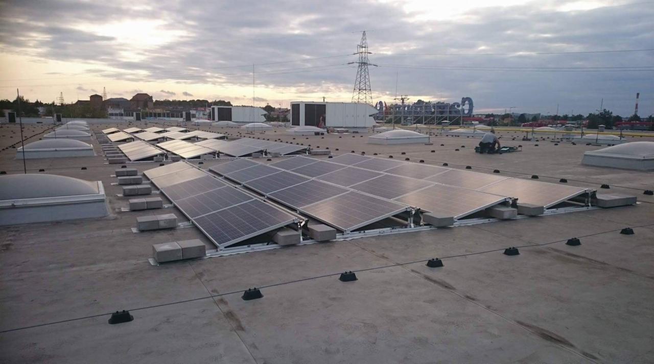 Panele słoneczne na płaskim dachu budynku komercyjnego, zamontowane na betonowych podstawach, z widocznym pracownikiem w trakcie serwisu, w tle słup wysokiego napięcia i zabudowania.