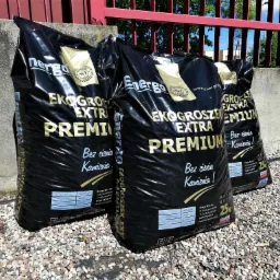 Trzy czarne worki z napisem 'EKOGROSZEK EXTRA PREMIUM' ustawione na żwirowym podłożu, na tle betonowego murku z czerwonym metalowym ogrodzeniem w tle.