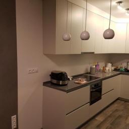 MebleWagerman - Nowoczesna, minimalistyczna kuchnia z białymi, matowymi frontami szafek, szarym blatem i trzema kulistymi lampami wiszącymi, widziana z boku, z widocznymi gniazdkami elektrycznymi na ścianie.