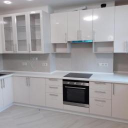 MebleWagerman - Biała, nowoczesna zabudowa kuchenna z błyszczącymi frontami i przeszklonymi szafkami wiszącymi, z piekarnikiem i płytą indukcyjną, częściowo jeszcze zabezpieczona folią ochronną.