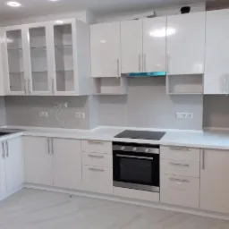 Biała, nowoczesna zabudowa kuchenna z błyszczącymi frontami i przeszklonymi szafkami wiszącymi, z piekarnikiem i płytą indukcyjną, częściowo jeszcze zabezpieczona folią ochronną.