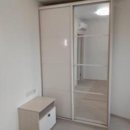 MebleWagerman - Jasna szafa przesuwna z lustrem w minimalistycznym wnętrzu, obok szafka nocna z otwartą półką i szufladą, widoczne oświetlenie sufitowe.