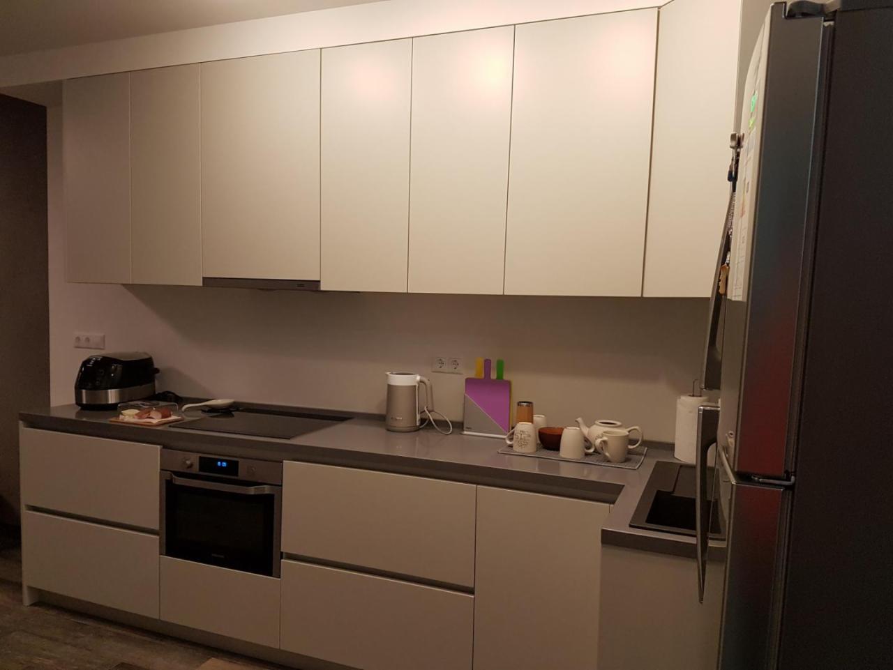 Nowoczesna kuchnia z matowymi, jasnoszarymi frontami szafek, szarym blatem i widocznym sprzętem AGD: piekarnik, toster i czajnik elektryczny, uzupełniona o zastawę do herbaty.