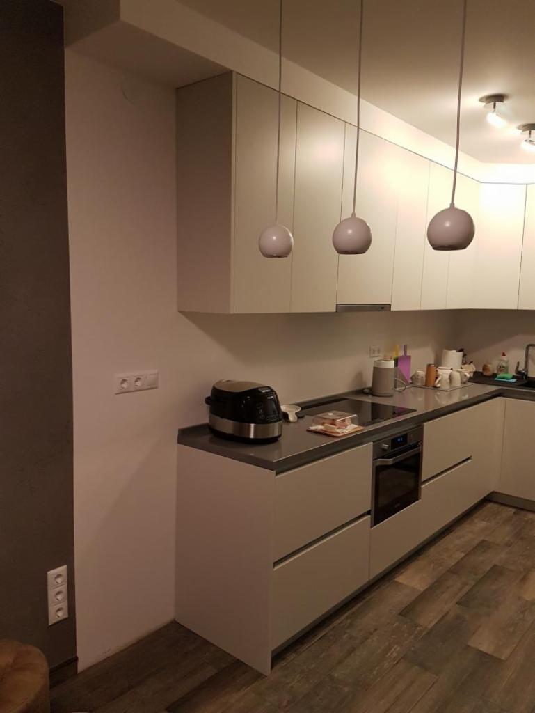 Nowoczesna, minimalistyczna kuchnia z białymi, matowymi frontami szafek, szarym blatem i trzema kulistymi lampami wiszącymi, widziana z boku, z widocznymi gniazdkami elektrycznymi na ścianie.