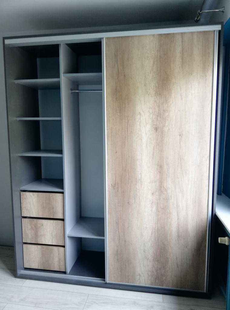 Szara szafa przesuwna z drewnopodobnymi drzwiami, widoczne półki i trzy szuflady w jasnym odcieniu drewna, wnętrze szafy w kolorze szarym.