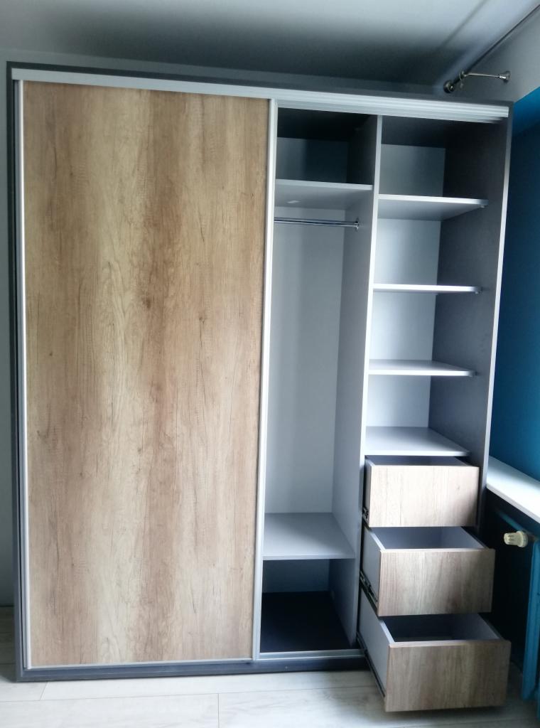 Nowoczesna szafa przesuwna z frontem w kolorze jasnego drewna i białym korpusem, widoczne półki i trzy szuflady w dolnej części.