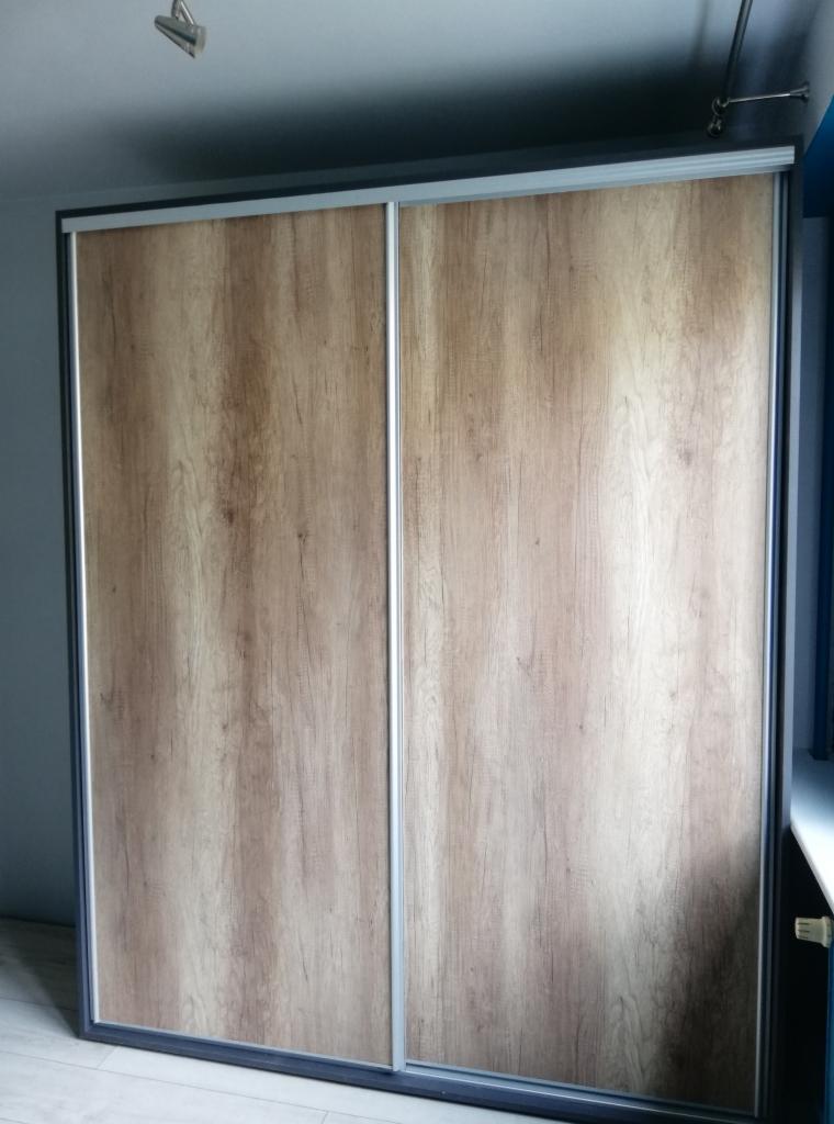 Dwudrzwiowa szafa przesuwna z okleiną imitującą jasne drewno, z szarymi ramami i srebrnymi prowadnicami, widoczna na tle szaro-niebieskiej ściany.