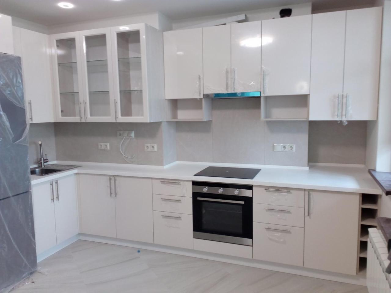 Biała, nowoczesna zabudowa kuchenna z błyszczącymi frontami i przeszklonymi szafkami wiszącymi, z piekarnikiem i płytą indukcyjną, częściowo jeszcze zabezpieczona folią ochronną.