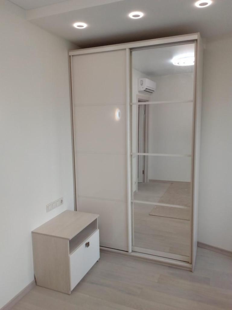 Jasna szafa przesuwna z lustrem w minimalistycznym wnętrzu, obok szafka nocna z otwartą półką i szufladą, widoczne oświetlenie sufitowe.