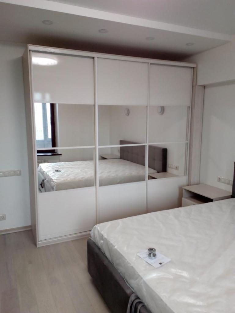 Biała szafa przesuwna z lustrzanymi drzwiami w sypialni, widoczne odbicie łóżka i stolika nocnego, laminowana podłoga, minimalistyczny design.