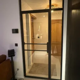 Szklane drzwi wewnętrzne z czarną ramą i poziomymi szprosami, widok na przedpokój z drewnianymi drzwiami i dywanikiem.