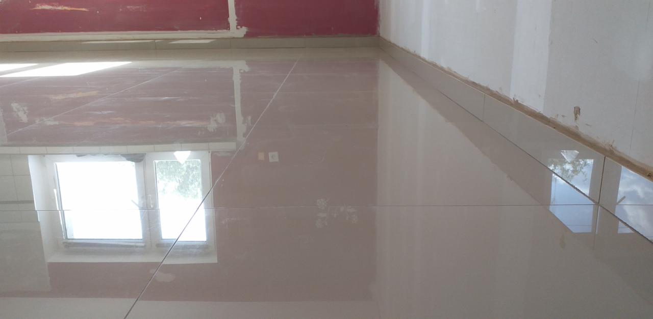 Świeżo ułożone, błyszczące płytki podłogowe w jasnym odcieniu beżu, z widocznym odbiciem okna i nieba, przy ścianie z widocznymi śladami prac wykończeniowych.