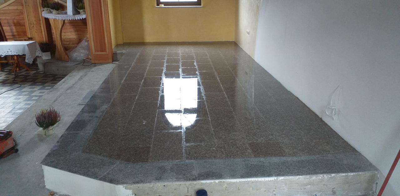 Świeżo ułożona, błyszcząca posadzka z płytek imitujących kamień, z widocznym odbiciem światła okiennego, na podwyższeniu w pomieszczeniu z żółtą ścianą i fragmentem drewnianej boazerii.