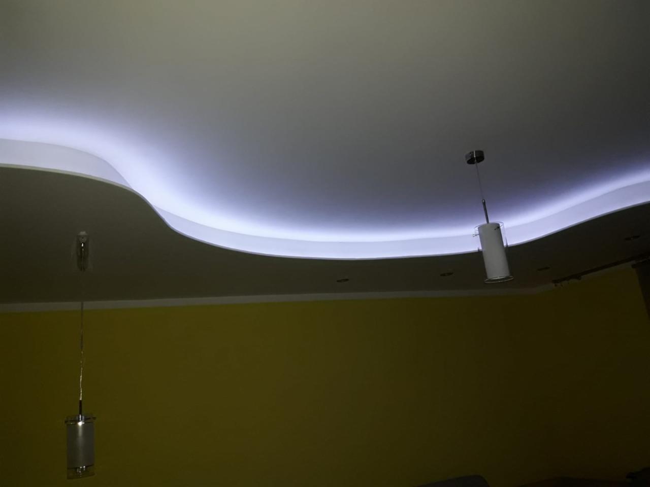 Podwieszany sufit z falistym obniżeniem i oświetleniem LED, widoczne dwie wiszące lampy cylindryczne na tle żółtej ściany.