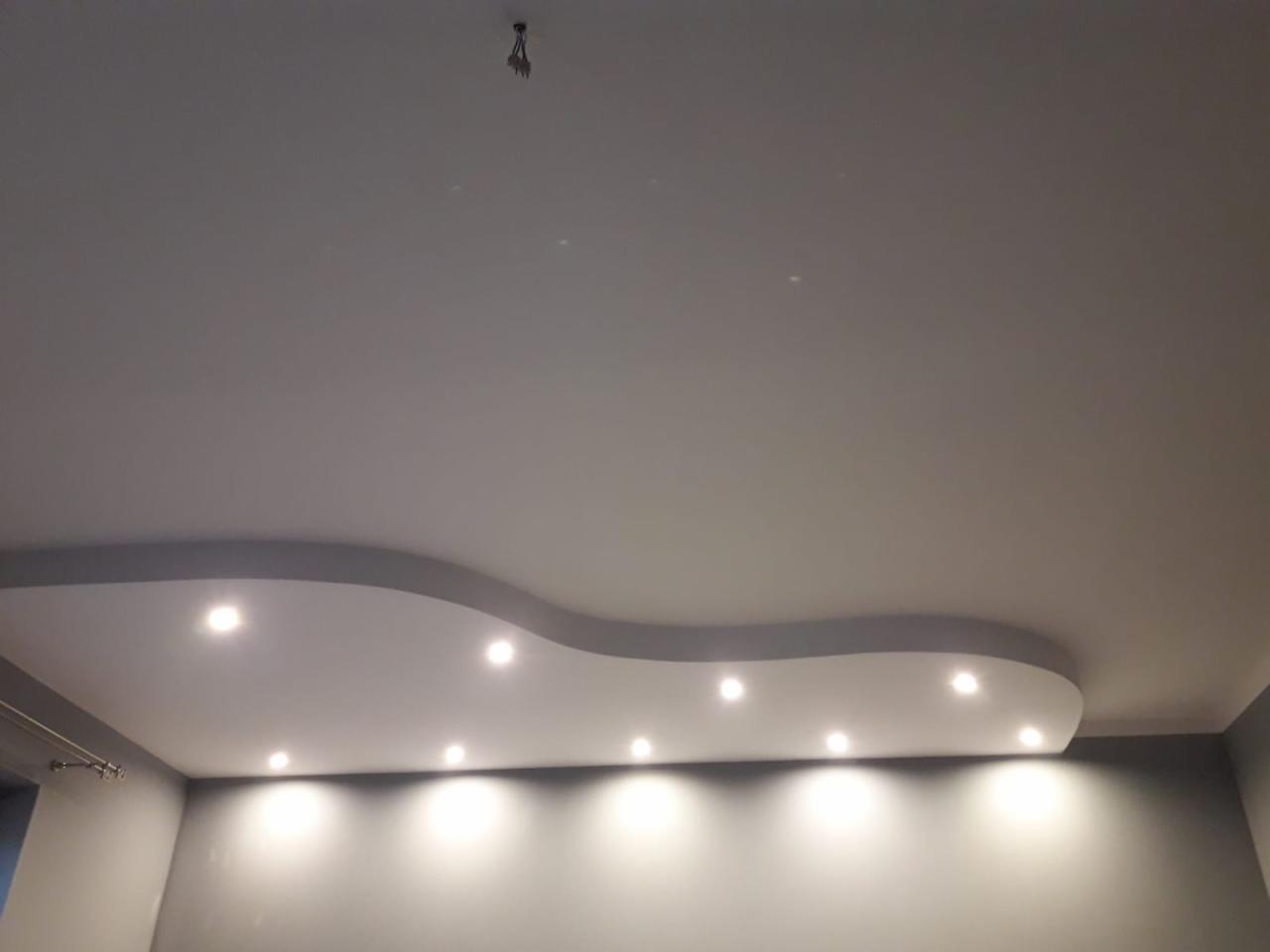 Gładki, szary sufit z dekoracyjnym podwieszanym elementem w kształcie fali, w którym zamontowano punktowe oświetlenie LED. Widoczny fragment ściany w tym samym odcieniu szarości.