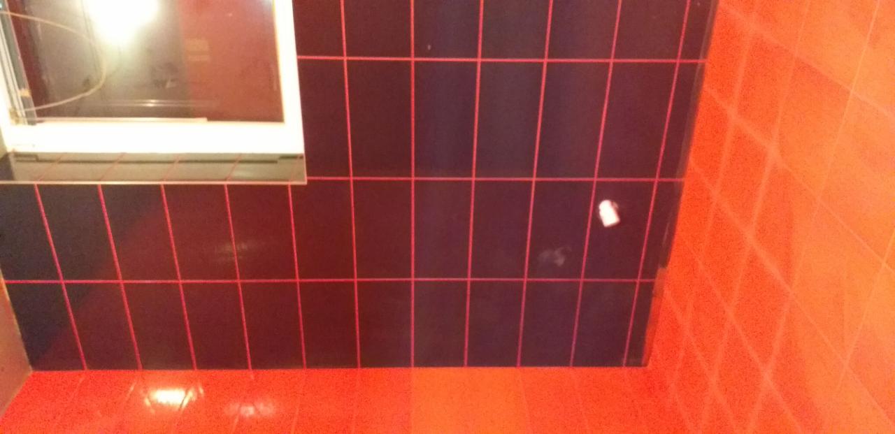 Ciemne, błyszczące płytki na ścianie z czerwoną fugą, kontrastujące z pomarańczową podłogą i fragmentem ściany, z widocznym oknem w górnej części kadru i drobnym zabrudzeniem na jednej z płytek.