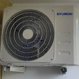 Zewnętrzna jednostka klimatyzacji Hyundai zamontowana na ścianie, widoczne wsporniki montażowe i niebieskie elementy wewnątrz wentylatora.