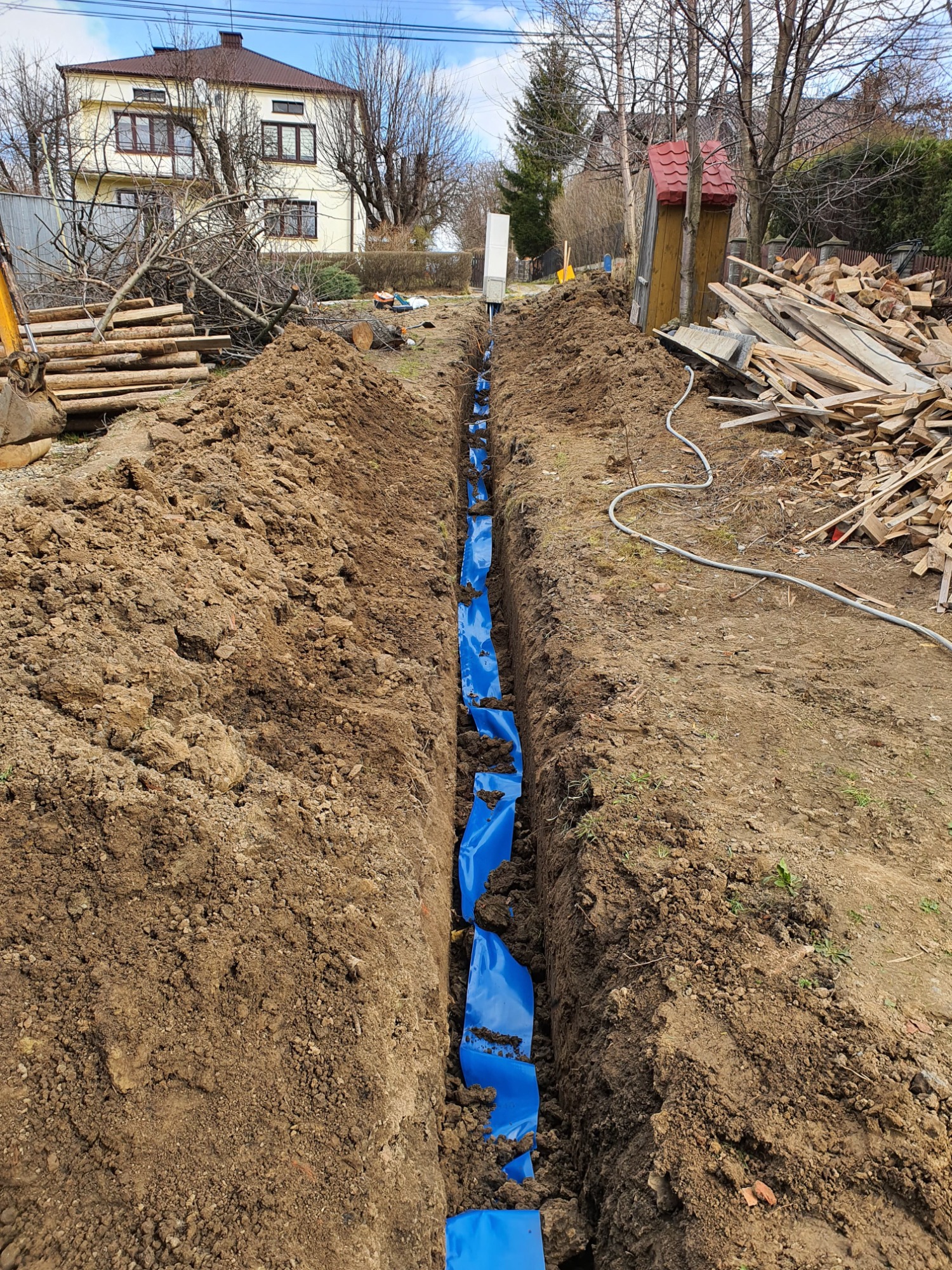 Wykopany rów z niebieską folią zabezpieczającą, przygotowany pod instalację przyłącza elektrycznego na działce z domem w tle.