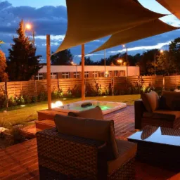 Oświetlony taras drewniany z jacuzzi, meblami ogrodowymi z rattanu i żaglami przeciwsłonecznymi o zmierzchu.